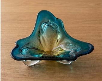 May include: A free-form glass bowl featuring a gradient color transition from a deep teal blue at the outer edges to a warm amber yellow in the center. The bowl has an organic, undulating rim with three distinct points and a smooth, polished finish.