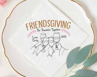 May include: A white Friendsgiving napkin on a plate with a gold rim. The napkin has the words "FRIENDSGIVING" and "Be Thankful Together" with a drawing of hands toasting wine glasses. The year 2025 is also printed on the napkin.