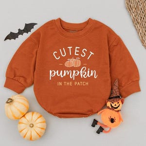 May include: An orange baby bodysuit with the text "Cutest Pumpkin in the Patch" in white. The image includes two small pumpkins, a bat cutout, and a Halloween-themed doll.