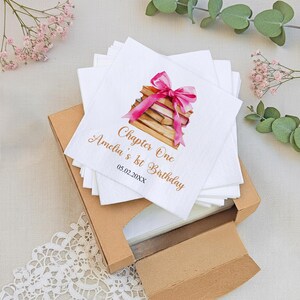 May include: White square napkins with a watercolor illustration of a stack of books tied with a pink ribbon. The napkins have gold text that reads "Chapter One Amelia's 1st Birthday 05.02.20XX". The napkins are in a brown cardboard box.