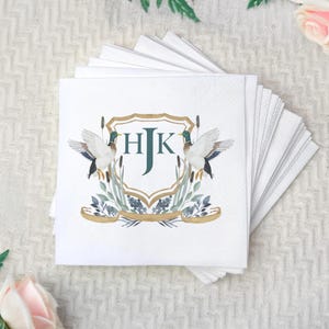 Può includere: Tovaglioli quadrati bianchi con un design decorativo. Il design presenta due anatre che fiancheggiano uno stemma con le iniziali "HJK" in verde. Lo stemma è circondato da canne e fogliame nei toni del blu, verde e oro. È visibile una pila di tovaglioli.