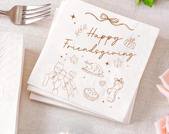 May include: White paper napkins with a brown illustration and the text "Happy Friendsgiving." The design features a turkey, wine glasses, and autumn leaves. A silver fork is visible.