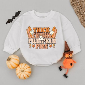 May include: White baby bodysuit with the text "Thick Thighs & Pumpkin Pies" in orange and pink. The bodysuit has long sleeves and a round neckline. Decorative pumpkins and a Halloween figure are next to the bodysuit.