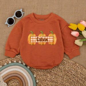 May include: Rust-colored baby romper with long sleeves. The front features three watercolor pumpkins with green stems and a plaid banner that reads "Calvin". A pair of blue sunglasses and a bouquet of flowers are next to the romper.