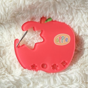May include: A bright red apple-shaped sewing needle holder with a bite taken out, revealing a star-shaped cutout. The word "apple" is written on a sticker. The holder has a green leaf and three flower-shaped cutouts.
