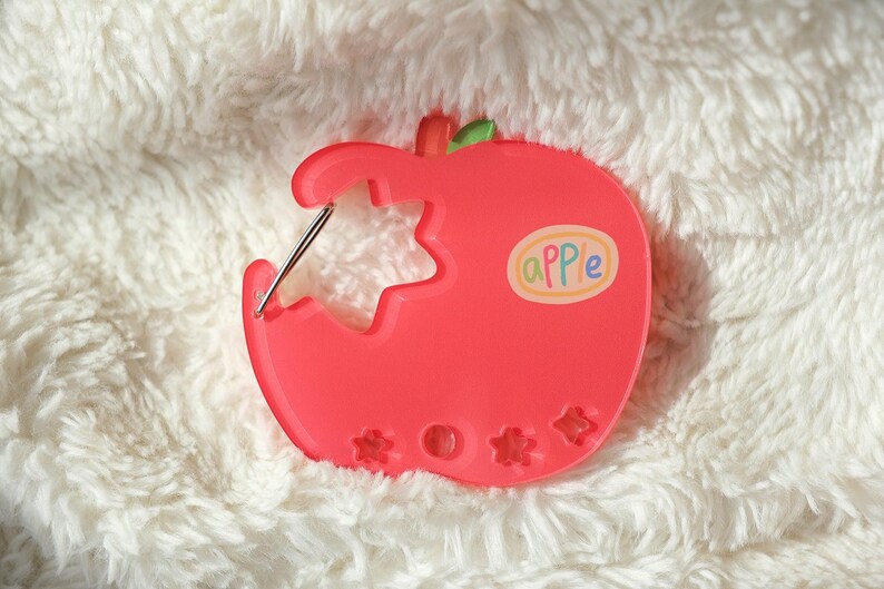 May include: A bright red apple-shaped sewing needle holder with a bite taken out, revealing a star-shaped cutout. The word "apple" is written on a sticker. The holder has a green leaf and three flower-shaped cutouts.