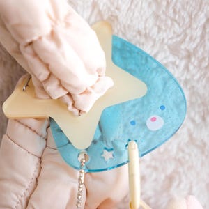 Op de afbeelding: Een babyspeeltje met een lichtblauw berenvormig element met een stervormig accent. Het speelgoed heeft een lichtgele ster en een kleine ketting. Het speelgoed wordt vastgehouden door een lichtroze gewatteerd item.