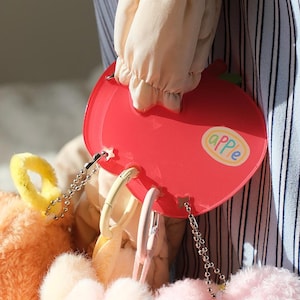 May include: A red apple-shaped accessory with the word "apple" printed on it. The accessory has several keychains attached to it. The background includes a person's sleeve and a plush toy.