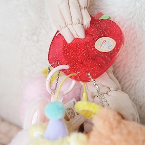 May include: A red, glittery apple-shaped keychain with the word "APPLE" on it. The keychain is attached to a chain and other colorful accessories, including a pink ring and a yellow cord. The background is a soft, light-colored fabric.