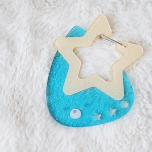 May include: A blue and cream-colored keychain with a star-shaped design. The keychain has a light blue base with a cream-colored star overlay and a silver clasp. The design includes small star and circle details.
