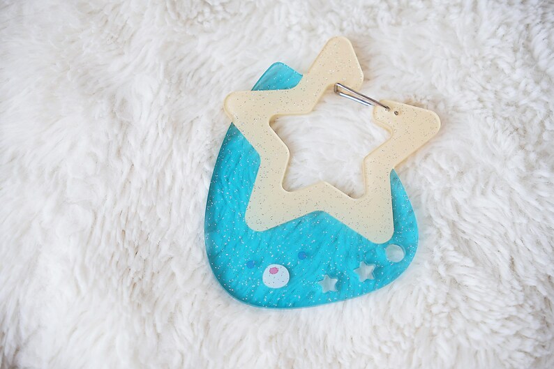 May include: A blue and cream-colored keychain with a star-shaped design. The keychain has a light blue base with a cream-colored star overlay and a silver clasp. The design includes small star and circle details.