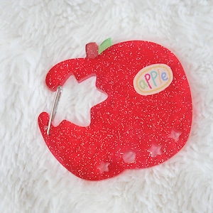 May include: A red glittery apple-shaped keychain with a bite taken out, revealing a star-shaped cutout. The keychain has a small sticker that says "apple". The keychain is on a white fluffy surface.