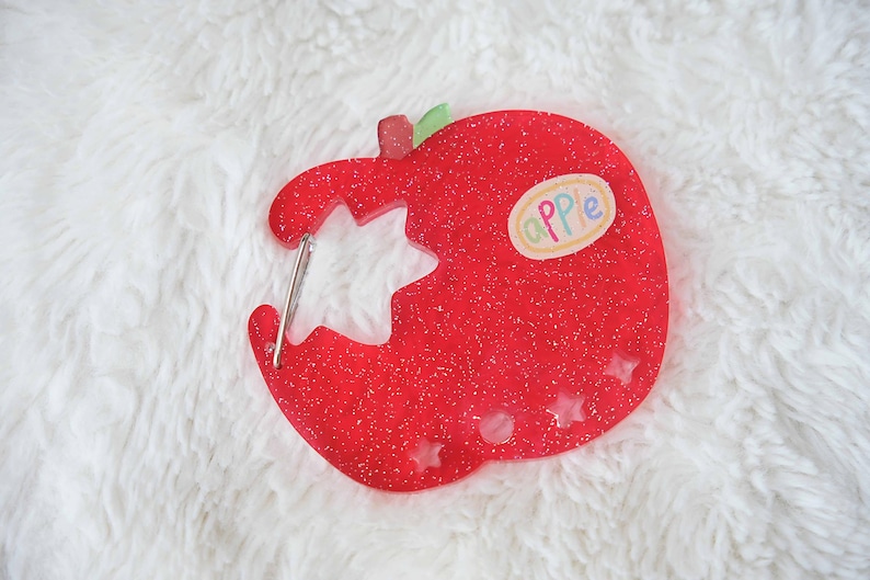 May include: A red glittery apple-shaped keychain with a bite taken out, revealing a star-shaped cutout. The keychain has a small sticker that says "apple". The keychain is on a white fluffy surface.