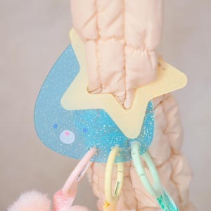 May include: A baby toy with a light blue, glittery teardrop shape and a yellow star. The toy has a pink and white face and is attached to a soft, quilted fabric. Colorful plastic rings hang from the toy.
