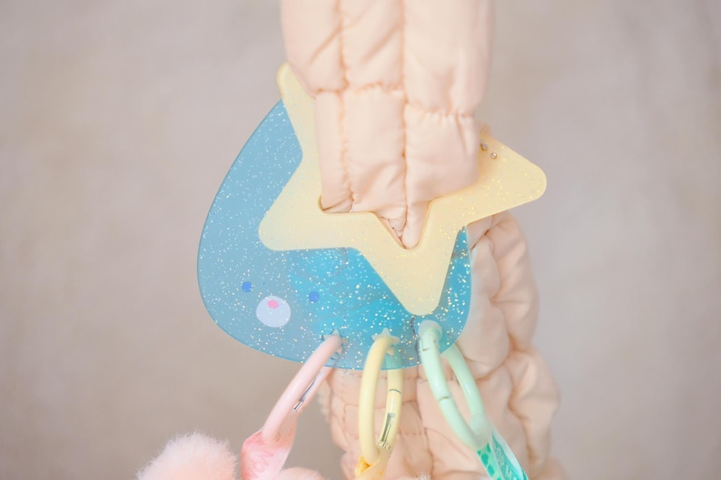 May include: A baby toy with a light blue, glittery teardrop shape and a yellow star. The toy has a pink and white face and is attached to a soft, quilted fabric. Colorful plastic rings hang from the toy.