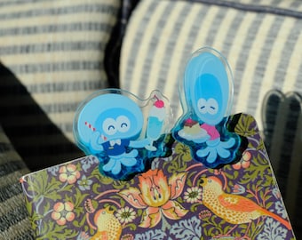 May include: Two blue jellyfish-shaped bookmarks with smiling faces, one holding a milkshake and the other a dessert, are placed on a patterned book. The book has a floral and bird design. The background is a striped pillow.