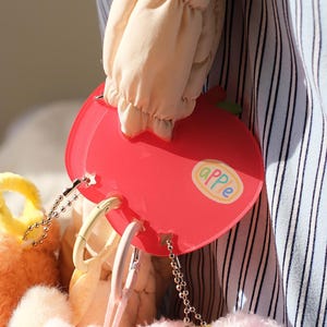 May include: A red apple-shaped accessory with the word "apple" on it. The accessory has a chain and is attached to a plush toy. The background includes a striped fabric and a light-colored sleeve.