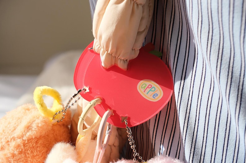 May include: A red apple-shaped accessory with the word "apple" on it. The accessory has a chain and is attached to a plush toy. The background includes a striped fabric and a light-colored sleeve.