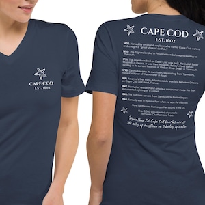 May include: Navy blue v-neck t-shirt with a white star and "CAPE COD EST. 1602" printed on the front. The back of the shirt features a list of historical facts about Cape Cod, with star graphics. The shirt is made of a soft material.