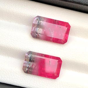 May include: Two rectangular watermelon tourmaline gemstones with a pink and green colour gradient. The gems are faceted with a rectangular shape and are displayed on a white surface.