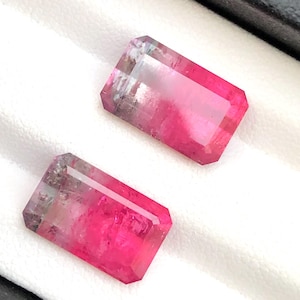 May include: Two rectangular gemstones with a faceted cut, displaying a vivid pink colour. The gems have a gradient effect, shifting from a lighter, translucent top to a deeper pink at the base. They are set against a white background.