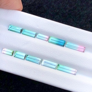 May include: Several rectangular gemstones with a colour gradient, including blue, green, and pink. The gems are displayed on a white surface, highlighting their polished facets. These are likely tourmaline gemstones, often used in jewellery.