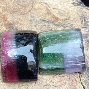 May include: Two rectangular gemstones with polished surfaces. One is a gradient of pink, black, and purple. The other is green and purple. The stones are displayed on a textured, brown surface.