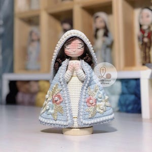 May include: A handmade crochet doll of a religious figure, with a light blue cloak featuring floral embroidery and a white dress. The doll has closed eyes and clasped hands, with a white beaded hood. The doll is on a wooden base.