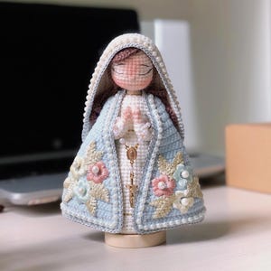 May include: A handmade crochet Virgin Mary figurine. It features a light blue cloak decorated with pink, white, and blue floral details. The figure has a white veil with pearl accents and a gold rosary. The figurine is mounted on a wooden base.