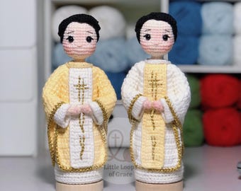 Może przedstawiać: Dwie ręcznie robione szydełkowe figurki postaci religijnych. Jedna postać ma na sobie żółto-białą szatę ze złotymi wykończeniami, druga białą szatę ze złotymi wykończeniami. Obie postacie mają czarne włosy i stoją na drewnianych podstawach. Postacie trzymają ręce razem w modlitwie.