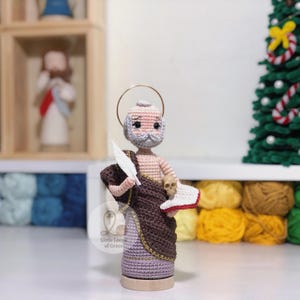 May include: Crocheted figure of a saint with a halo, holding a quill and a book. The figure is dressed in a brown and purple robe with gold trim. A small skull is next to the book. The figure is on a wooden base. The text "Little Loops of Grace" is visible.