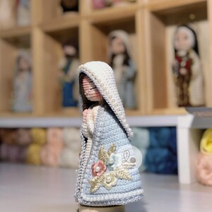 May include: A handcrafted crochet figure of a religious icon, with a light blue cloak adorned with floral details and a white head covering. The figure is presented on a wooden base, with similar figures visible in the background.
