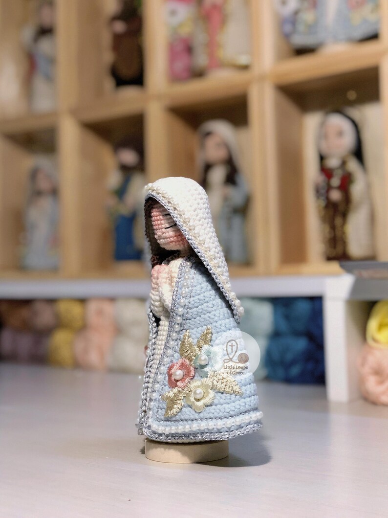 May include: A handcrafted crochet figure of a religious icon, with a light blue cloak adorned with floral details and a white head covering. The figure is presented on a wooden base, with similar figures visible in the background.