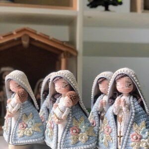 May include: Handmade crochet figurines of the Virgin Mary holding baby Jesus. The figures wear light blue cloaks with floral embroidery and pearl accents. The dolls have white veils and are on wooden bases.