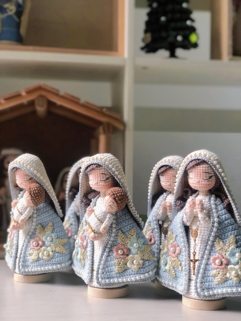 May include: Handmade crochet figurines of the Virgin Mary holding baby Jesus. The figures wear light blue cloaks with floral embroidery and pearl accents. The dolls have white veils and are on wooden bases.