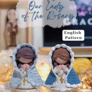 May include: Two crocheted figurines of Our Lady of the Rosary with brown hair, wearing white dresses and capes in light blue and dark blue. Each figure has a gold-colored cross pendant and hands clasped in prayer. Text reads Our Lady of the Rosary.