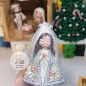 May include: A handmade crochet doll of a religious figure, wearing a light blue cloak with floral and pearl embellishments. The doll has a white veil and is held in a sheer bag. The doll is part of a nativity scene.