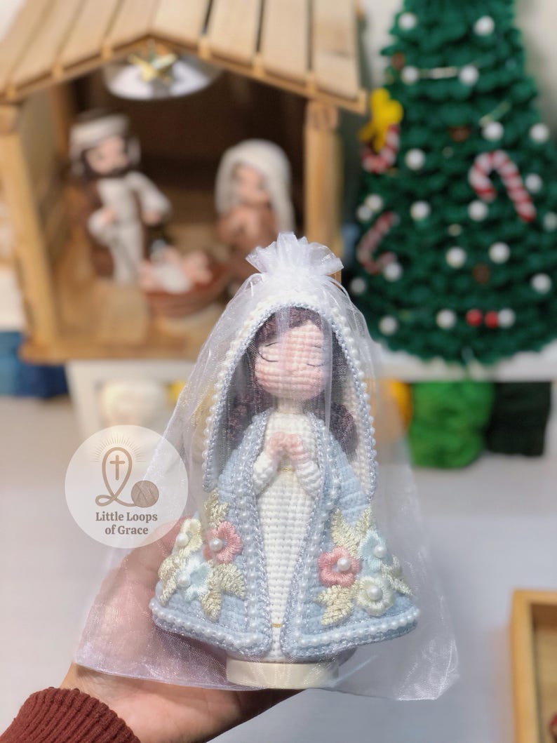 May include: A handmade crochet doll of a religious figure, wearing a light blue cloak with floral and pearl embellishments. The doll has a white veil and is held in a sheer bag. The doll is part of a nativity scene.