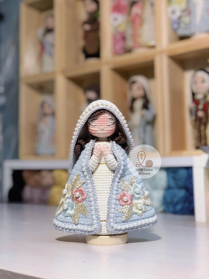 May include: A handmade crochet doll of a religious figure, with a light blue cloak featuring floral embroidery and a white dress. The doll has closed eyes and clasped hands, with a white beaded hood. The doll is on a wooden base.