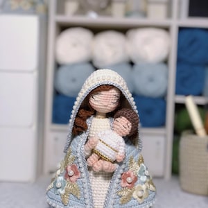 May include: Crocheted doll depicting a religious figure holding a baby. The doll has a white and light blue cloak with floral and pearl embellishments. The doll's hood is trimmed with pearls. The doll is on a wooden base.