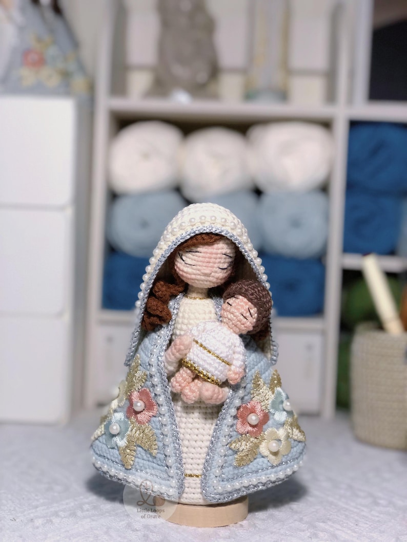 May include: Crocheted doll depicting a religious figure holding a baby. The doll has a white and light blue cloak with floral and pearl embellishments. The doll's hood is trimmed with pearls. The doll is on a wooden base.
