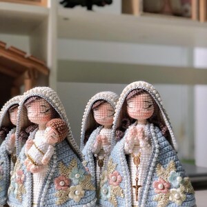May include: Crocheted religious figurines, featuring a light blue cloak adorned with floral embroidery and pearl accents. The figurines have closed eyes and are holding a baby or praying. The dolls are on wooden bases.