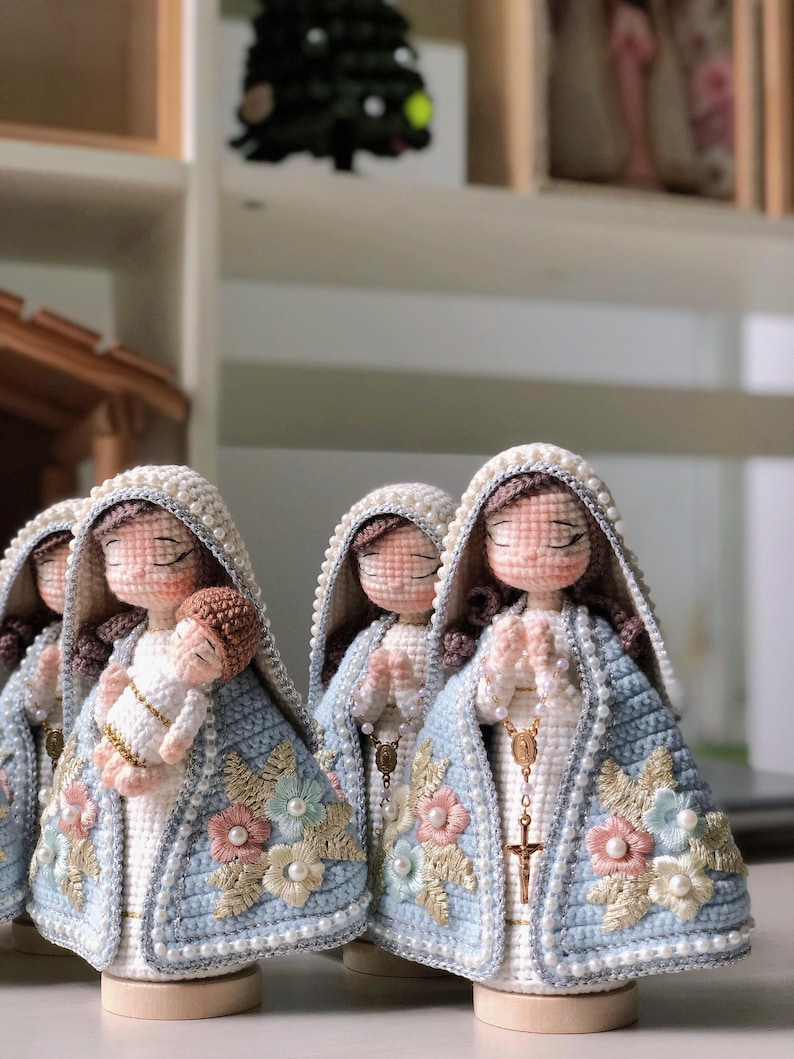 May include: Crocheted religious figurines, featuring a light blue cloak adorned with floral embroidery and pearl accents. The figurines have closed eyes and are holding a baby or praying. The dolls are on wooden bases.