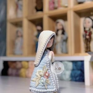 May include: A handcrafted crochet figurine of a religious figure, with a light blue dress decorated with floral details. The figure has a white head covering and clasped hands. Other similar figurines are visible in the background.