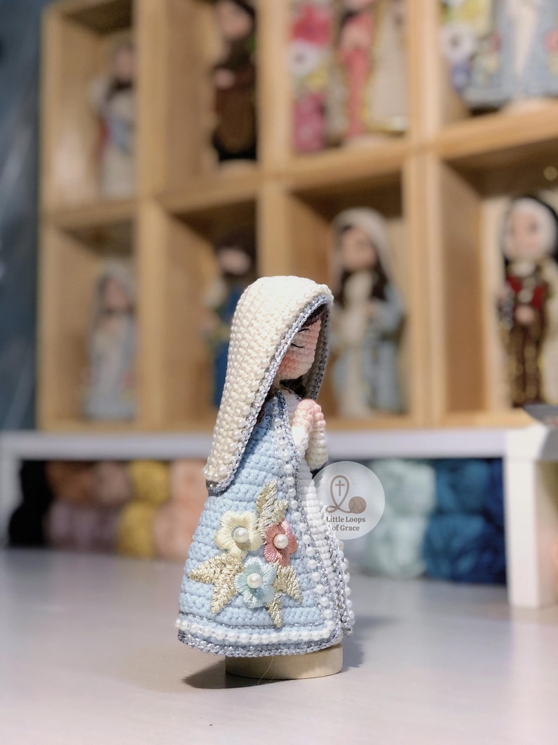 May include: A handcrafted crochet figurine of a religious figure, with a light blue dress decorated with floral details. The figure has a white head covering and clasped hands. Other similar figurines are visible in the background.
