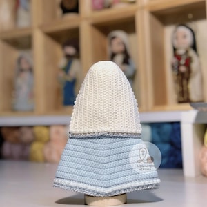 May include: A handmade, crocheted religious figurine with a white head covering and a light blue dress. The dress has a chevron pattern and is trimmed with silver yarn. The figurine is displayed on a wooden base.