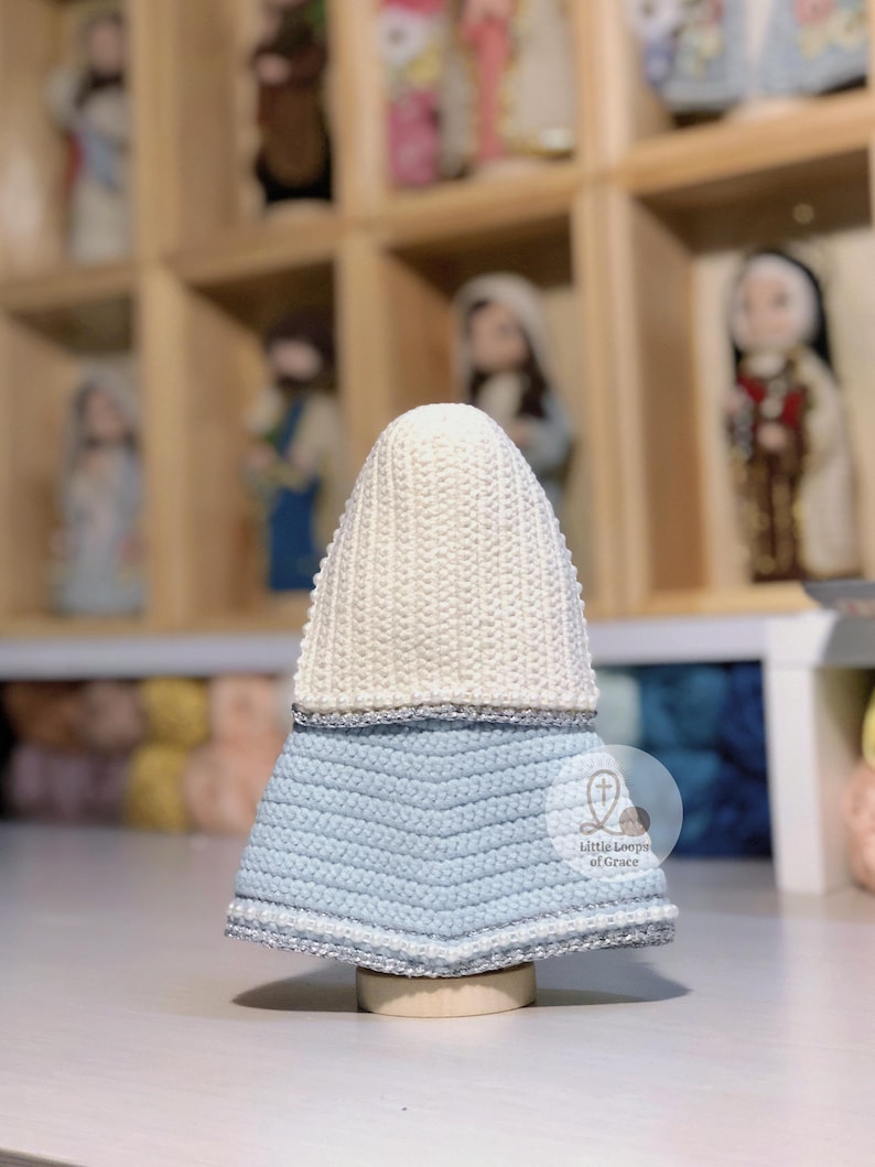 May include: A handmade, crocheted religious figurine with a white head covering and a light blue dress. The dress has a chevron pattern and is trimmed with silver yarn. The figurine is displayed on a wooden base.