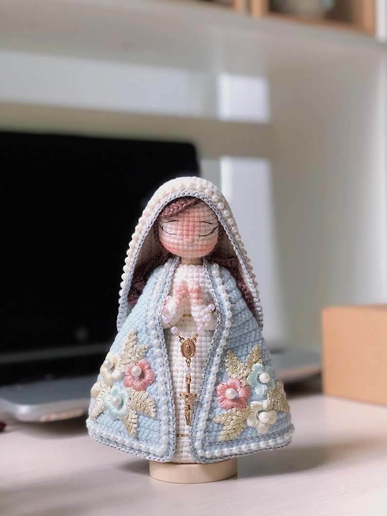 May include: A handmade crochet Virgin Mary figurine. It features a light blue cloak decorated with pink, white, and blue floral details. The figure has a white veil with pearl accents and a gold rosary. The figurine is mounted on a wooden base.