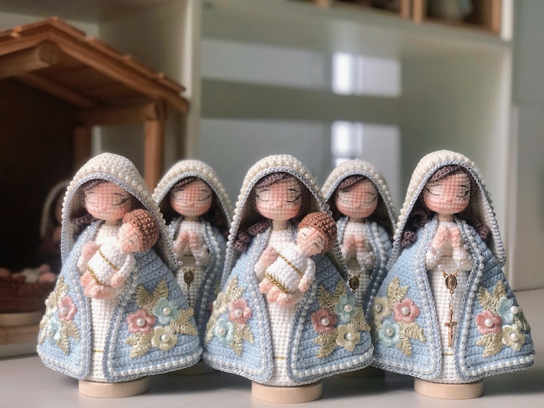 May include: Five handmade crochet dolls, each representing a religious figure holding a baby. The dolls are dressed in light blue and white robes, adorned with floral and pearl details. Each has a white head covering and closed eyes.