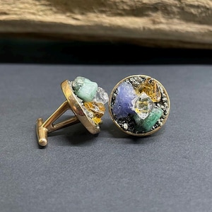 May include: A pair of gold-toned cufflinks, each featuring a cluster of colorful gemstones. The stones include green, blue, yellow, and clear crystals set against a dark, textured background. The cufflinks have a classic, elegant design.
